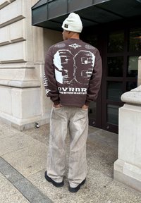 Άνδρας με λευκό σκουφάκι, καφέ φούτερ με μεγάλο λευκό αριθμό "96" και κείμενο στην πλάτη, μπεζ παντελόνι, στέκεται έξω κοντά στην είσοδο πέτρινης οικοδομής.