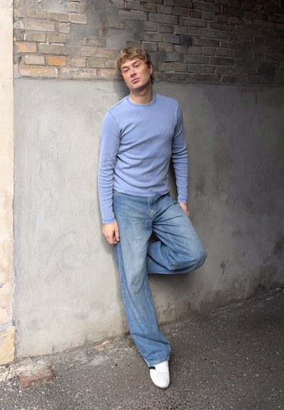 Joven con suéter azul claro y vaqueros se apoya en una pared bicolor de ladrillo y hormigón con una rodilla doblada, llevando zapatos blancos.