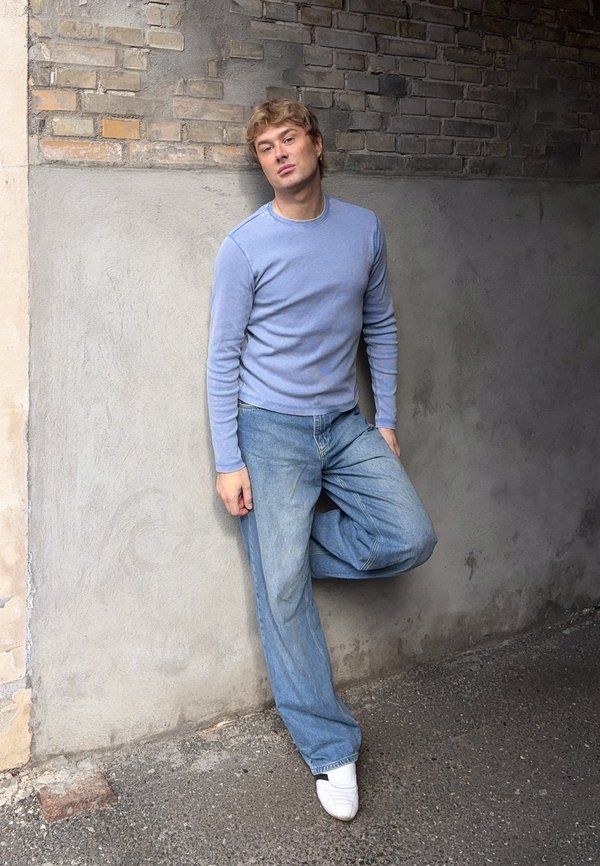Jauns vīrietis gaiši zilā džemperī un džinsās atspiežas pret divtoņu ķieģeļu un betona sienu ar vienu saliektu ceļgalu, valkājot baltas kurpes.