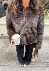 Giacca in pelliccia sintetica marrone con un ampio colletto, abbinata a pantaloni neri. Tiene in mano una tazza con una cannuccia e una borsa a fantasia. Sfondo in cemento.