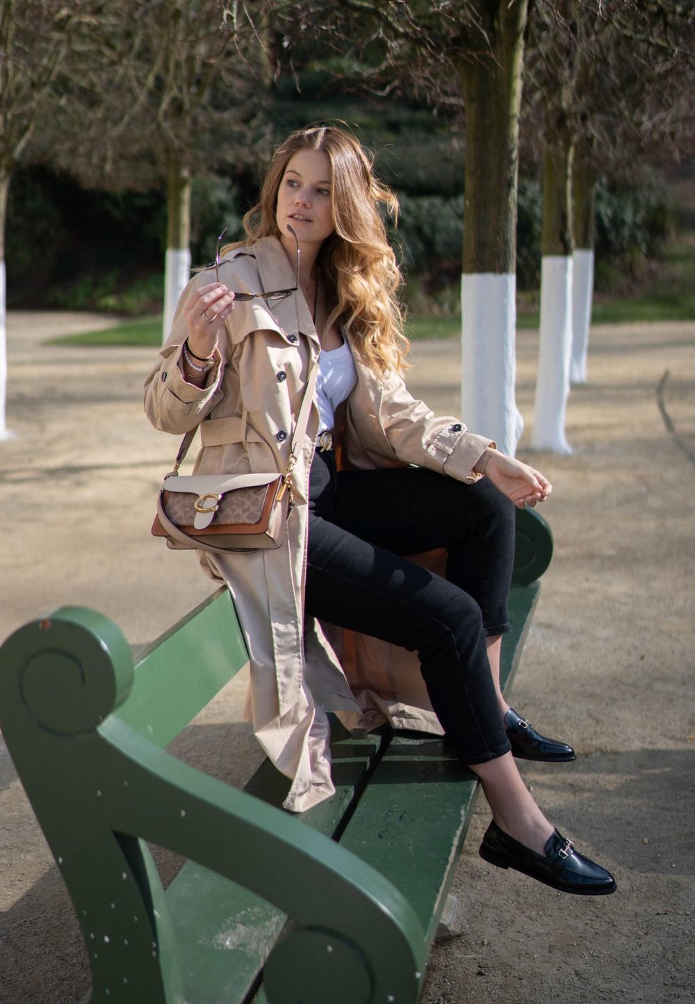 Casaco trench bege com cintura cingida, calças skinny pretas e mocassins pretos. Asegurando uns óculos de sol, sentado num banco verde.