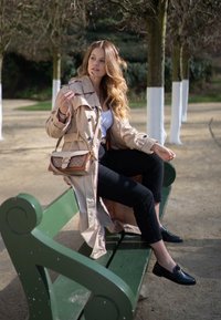 Casaco trench bege com cintura cingida, calças skinny pretas e mocassins pretos. Asegurando uns óculos de sol, sentado num banco verde.