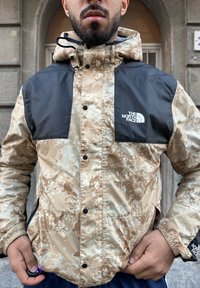 Beige and black waterproof jacket with a splatter pattern, zippered pockets, and logo on the chest, featuring a hood and elastic cuffs.