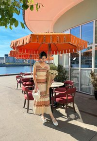 Μάξι φόρεμα με ζιγκ-ζαγκ ρίγες σε μπεζ, πορτοκαλί και καφέ. Λευκά παπούτσια, κρατώντας ένα μπουκέτο. Υπαίθριο σκηνικό κάτω από μια πορτοκαλί ομπρέλα.