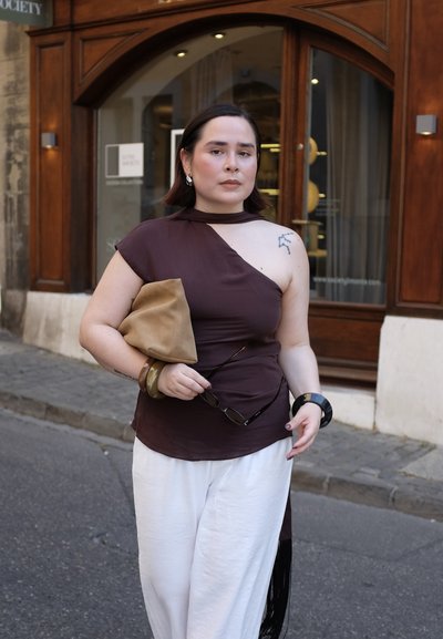 Top asimétrico marrón con cuello, combinado con pantalones de pierna ancha blancos. Sosteniendo un bolso beige y gafas de sol negras. Calle de concreto de fondo.