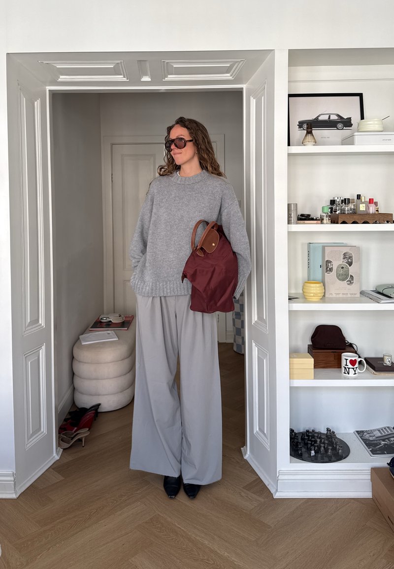 Femme en pull gris oversize et pantalon large tenant un sac bordeaux, debout dans l'embrasure d'une porte, à côté d'étagères blanches avec des livres, de la décoration et un jeu d'échecs.