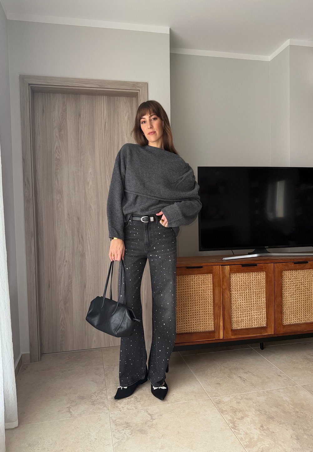 Kvinne som står innendørs iført en grå, oversized genser, svart jeans med nagler, svarte spisse sko, og holder en svart håndveske.