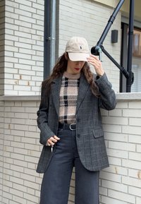 Hall triibuline villane bleiser beežide ja mustade ruutudega kampsuni kohal, täiendatud mustade laiade pükste ja kreemja pesapalli mütsiga.