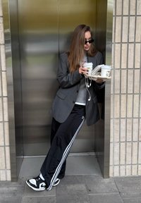 Person, der einen dunklen übergroßen Blazer über einem grauen Pullover und schwarzen Sporthosen mit weißen Streifen trägt, hält zwei Kaffeetassen und ein Tablett.