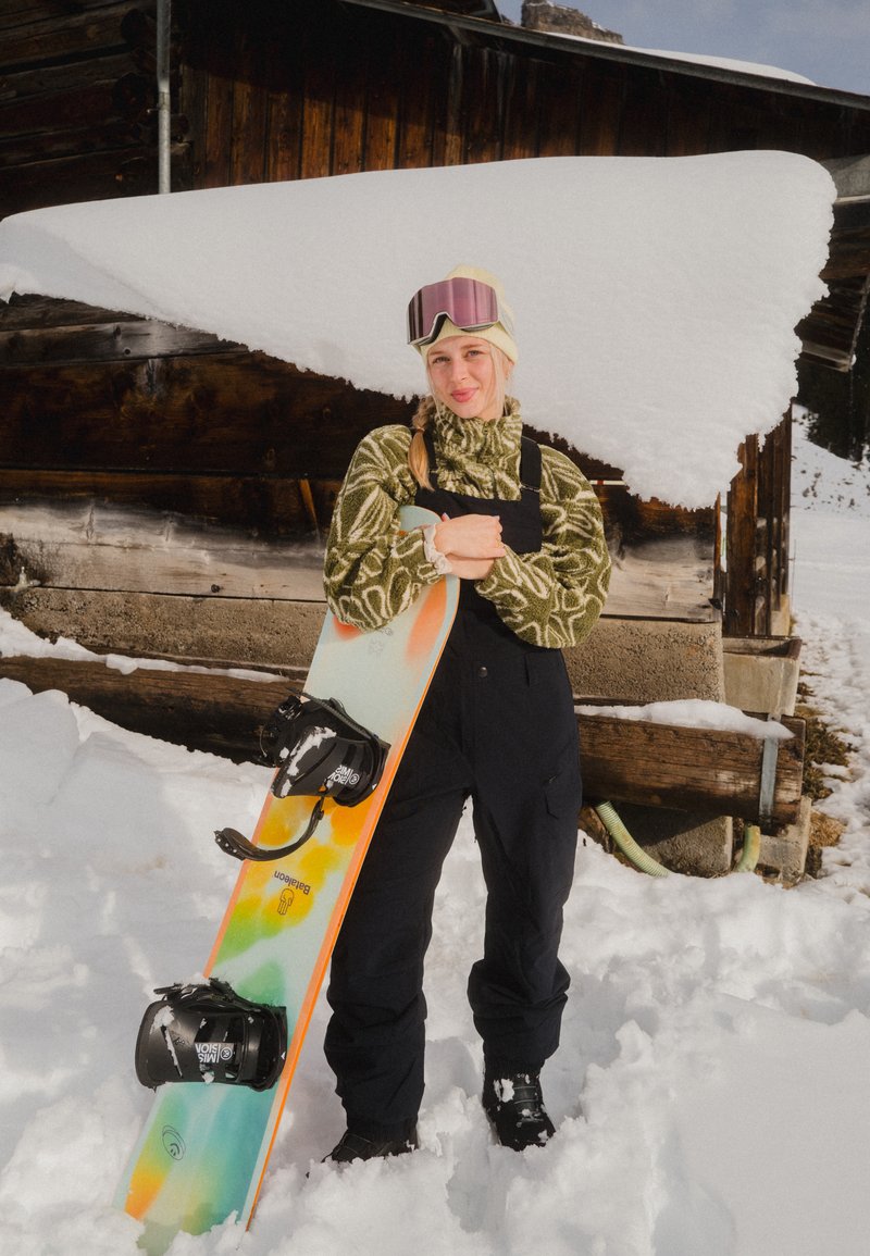 Jeune femme en tenue d'hiver tenant un snowboard coloré, debout dans la neige devant une cabane en bois avec neige sur le toit.