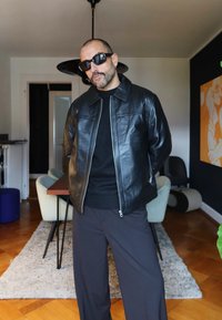 Svart skinnjakke med glidelås over en svart genser, kombinert med mørke bukser. Rommet har tregulv, spisestuebord og minimalistisk innredning.