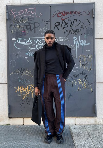 Una persona que lleva una camisa de manga larga negra, una chaqueta negra abierta y pantalones marrón con un patrón y rayas azules, sosteniendo un bolso negro.