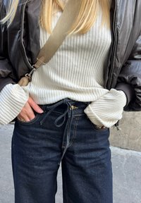 Ριχτό λευκό μπλούζα με μακριά μανίκια και φαρδιά μανσέττα, συνδυασμένο με σκούρα τζιν παντελόνια που έχουν μέση με κορδόνι και χρυσές ραφές.