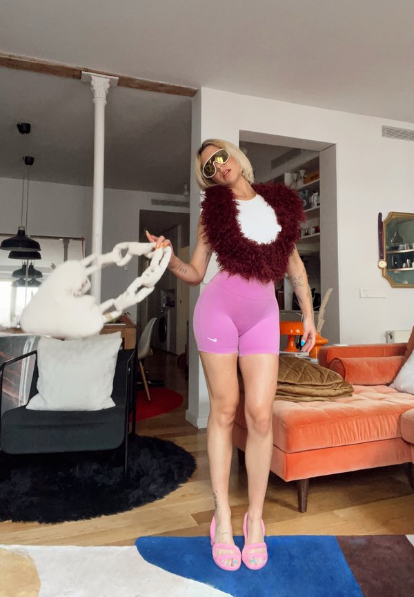 Žena oblečená v ružových šortkách, bielom topu, bordovej vesty s perím, slnečných okuliaroch a ružových lodičkách šúcha bielu kabelku v tvare reťaze v modernom obývacom priestore.