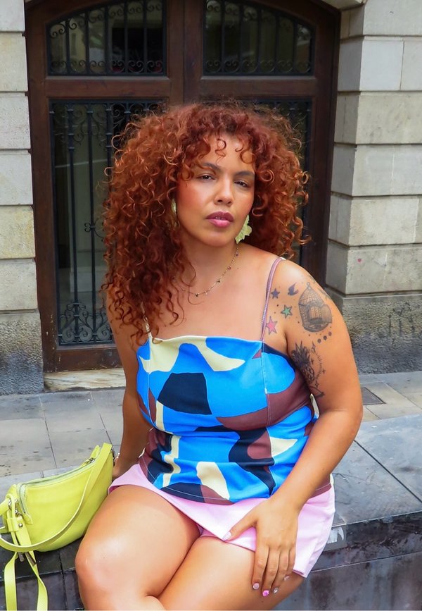 Femme aux cheveux roux bouclés et au bras tatoué coloré, portant un haut bleu à motifs et une jupe rose, assise sur un banc en pierre avec un sac jaune à côté d'elle.
