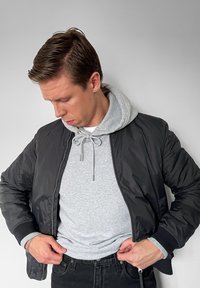 Czarna kurtka typu bomber o gładkiej fakturze, pod nią szara bluza z kapturem z troczkiem, czarne dżinsy oraz jasnoszary detal mankietu.