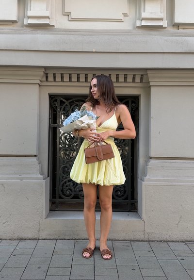 Vestido amarillo a la altura de la rodilla con tirantes finos; tejido texturizado. Sosteniendo un ramo de flores azules y un bolso marrón contra una pared decorativa.