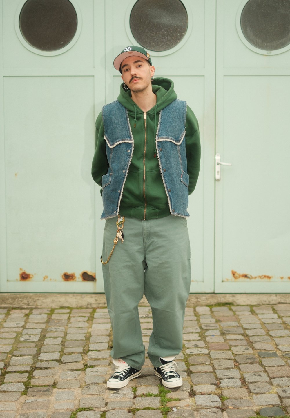 Veste en denim, sweat à capuche vert, et pantalon vert à jambes larges ; porte des baskets noires et blanches. Accessoires comprenant un porte-clés sur le pantalon. En arrière-plan, une porte vert menthe.