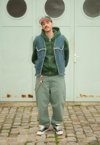 Veste en denim, sweat à capuche vert, et pantalon vert à jambes larges ; porte des baskets noires et blanches. Accessoires comprenant un porte-clés sur le pantalon. En arrière-plan, une porte vert menthe.