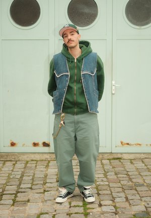 Veste en denim, sweat à capuche vert, et pantalon vert à jambes larges ; porte des baskets noires et blanches. Accessoires comprenant un porte-clés sur le pantalon. En arrière-plan, une porte vert menthe.