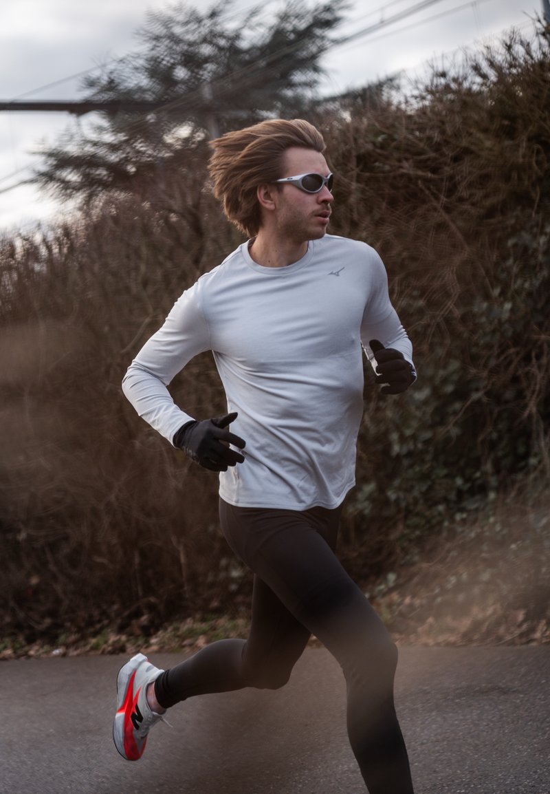Vyras su akiniais nuo saulės, pilka ilgomis rankovėmis marškinėliais, juodomis tamprėmis ir pirštinėmis bėga lauke asfaltuotu taku, fone krūmai.