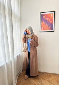 Lang beige frakk med svarte knapper, blå genser og lys grå skjerf. Rom med hvite gardiner og en innrammet fargerik grafisk utskrift.