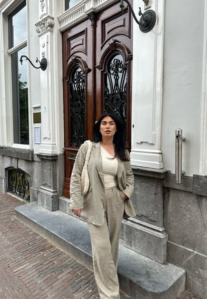 Traje de lino beige con una chaqueta de corte holgado y pantalones, combinado con una blusa color crema. El fondo presenta puertas dobles de madera y escaleras de piedra.