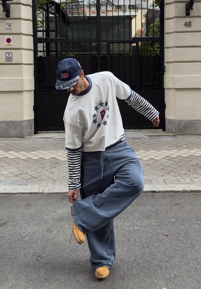 Hombre con camisa de rayas y gorra atándose una bota amarilla en una calle de la ciudad frente a una puerta negra y un edificio beige.