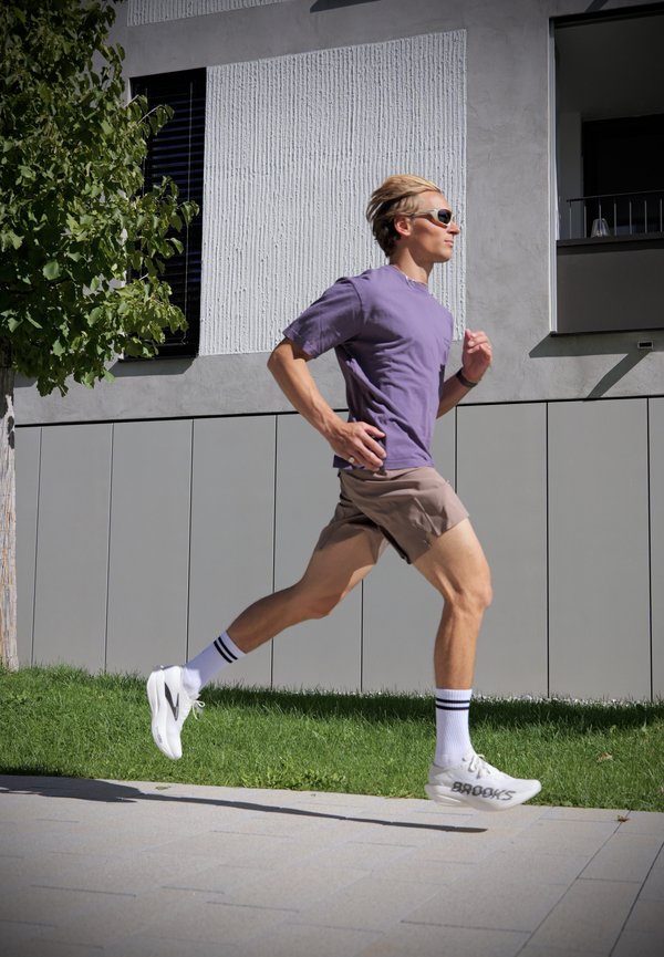 Un hombre con gafas de sol, una camiseta morada y ropa de running trota por una acera junto a un edificio moderno.