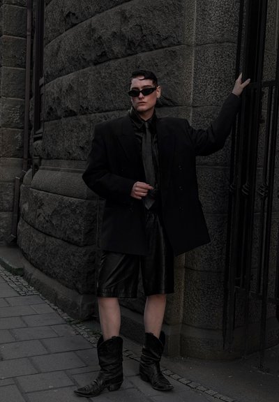 Blazer oversize de cuero negro, pantalones cortos a juego y botas de vaquero negras. Los accesorios incluyen gafas de sol elegantes y una corbata oscura, todo ello frente a una pared de piedra.