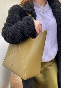 Olivovo zelená kožená taška s pevnou konštrukciou, špicatými okrajmi, bielym prešívaním a zlatým logom, ktorú drží osoba v čiernom huňatom kabáte.