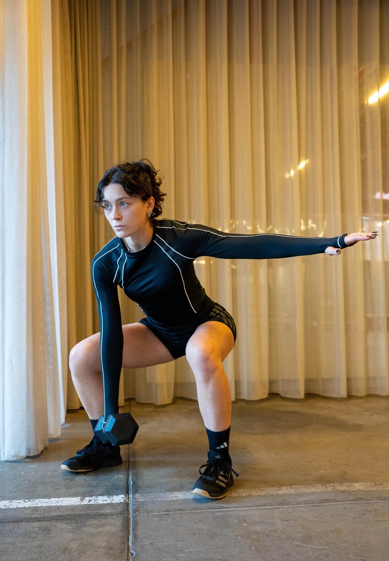 Femeie îmbrăcată în echipament de fitness negru face genuflexiuni în interior, ținând o ganteră într-o mână și cu cealaltă braț întins pentru echilibru.