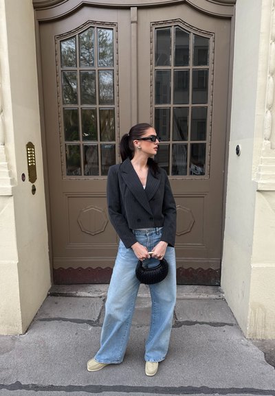 Mujer con gafas de sol, vistiendo un blazer oscuro corto, jeans de pierna ancha en azul claro, zapatos crema, sosteniendo un bolso de mano de mimbre negro, de pie junto a unas puertas dobles marrones.