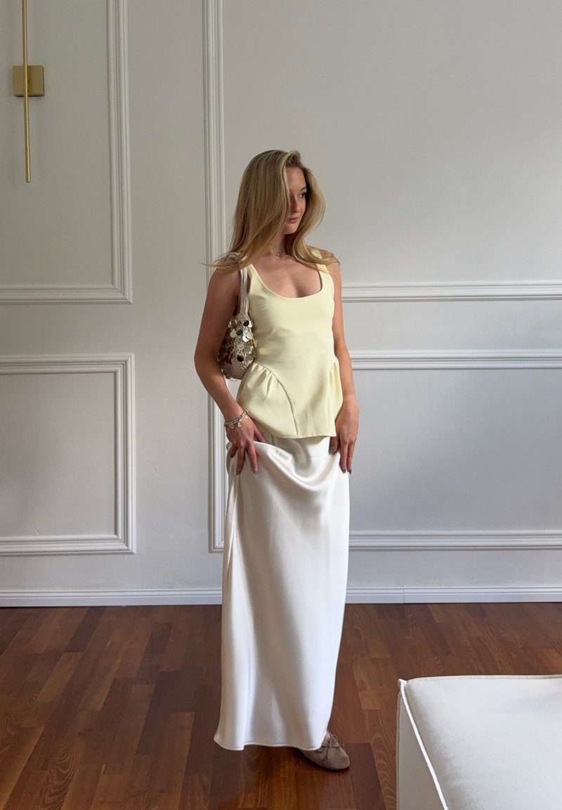 Femme blonde portant un haut sans manches jaune pâle, une longue jupe blanche, des chaussures marron et tenant un petit sac à main beige, debout sur un sol en bois à l'intérieur.