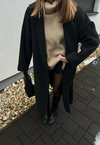 Personne portant un long manteau noir, un pull à col roulé côtelé beige, un short noir, des bottes noires et tenant un sac à main noir, debout sur un chemin pavé.