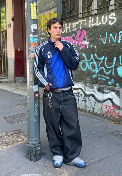 Chaqueta de entrenamiento azul y negra de Adidas con rayas, jeans de mezclilla oscura holgados y zapatos grises. Llavero visible en la cintura, de pie cerca de una pared con graffiti.