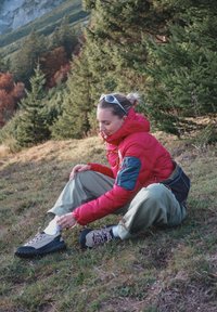 Žena v červené bundě zavazuje turistické boty, zatímco sedí na trávě s borovicemi a horami v pozadí během podzimu.