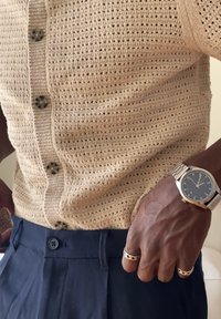 Sötét bőrű személy bézs színű kötött, gombos felsőt, tengerészkék nadrágot, ezüstszínű karórát és két ezüst mintás gyűrűt visel a jobb kezén.