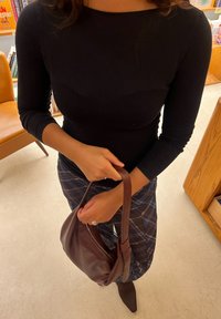 A person wearing a black long-sleeve top and a plaid skirt is holding a brown leather handbag in a room furnished with wooden furniture and bookshelves.
