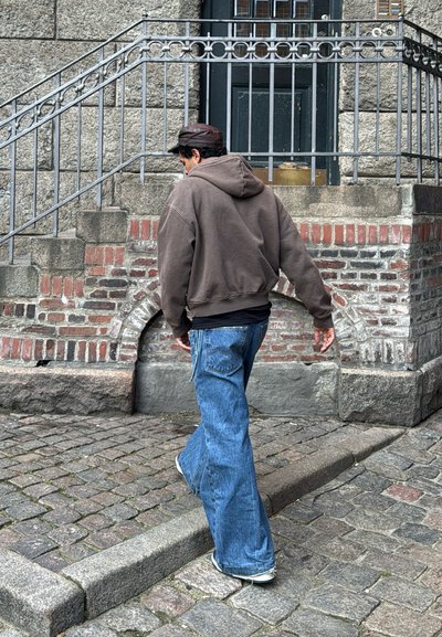Sudadera con capucha marrón, pantalones de mezclilla acampanados azules, a lo largo de una calle adoquinada cerca de un muro de piedra, subiendo escaleras de piedra con barandilla de hierro.