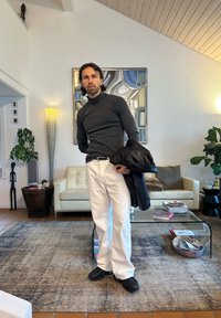 Czarny sweter z golfem, białe spodnie o prostych nogawkach oraz czarne buty; stojący w nowoczesnym salonie z beżową sofą i dywanem.