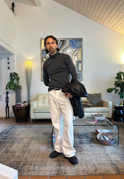 Jersey de cuello alto negro, pantalones rectos blancos y zapatos negros; de pie en una sala de estar moderna con un sofá y alfombra beige.