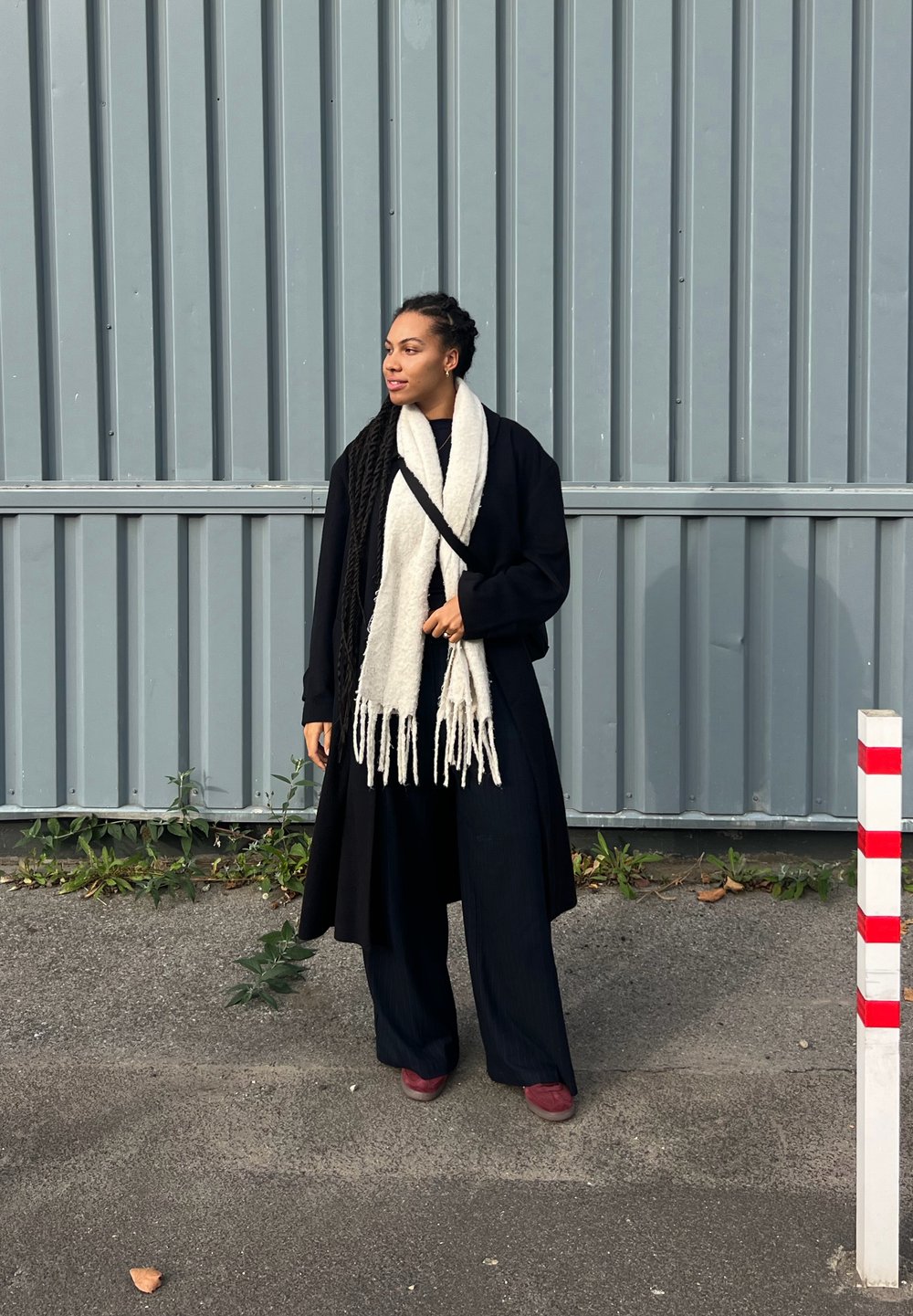 Crni dugi kaput preko tamne odjeće, s debelim bijelim šal na kojem su rese, stoji ispred sive pozadine s okomitim panelima.