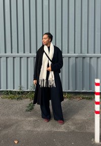 Crni dugi kaput preko tamne odjeće, s debelim bijelim šal na kojem su rese, stoji ispred sive pozadine s okomitim panelima.