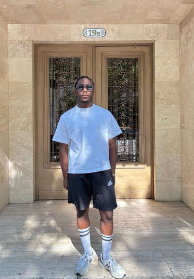 Camiseta blanca de manga corta, pantalones cortos negros con detalles blancos, calcetines blancos hasta la rodilla, zapatillas grises; de pie frente a puertas de madera ornamentadas.