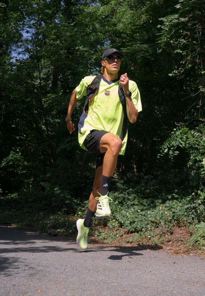 Camiseta deportiva de color amarillo brillante, pantalones cortos negros y calcetines negros. Las zapatillas de correr son de color gris claro con cordones verdes. El fondo presenta vegetación y un camino.