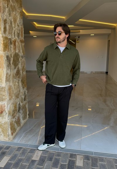 Suéter verde oliva, tejido texturizado, con cuello, superpuesto sobre una camisa blanca. Pantalones anchos negros y zapatillas blancas con detalles verdes.