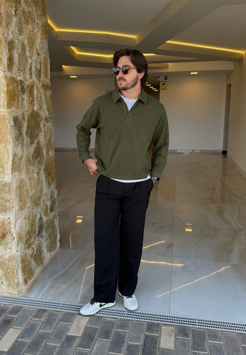 Olijfgroen pullover, gestructureerde stof, met kraag, laag over een wit shirt. Zwarte wijde broeken en witte sneakers met groene accenten.