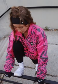 Rožinė ir pilka vėjo nepraleidžianti striukė su užtrauktuku, juodos tamprės ir baltos sportbačiai atliekant pritūpimo pozą ant laiptų.