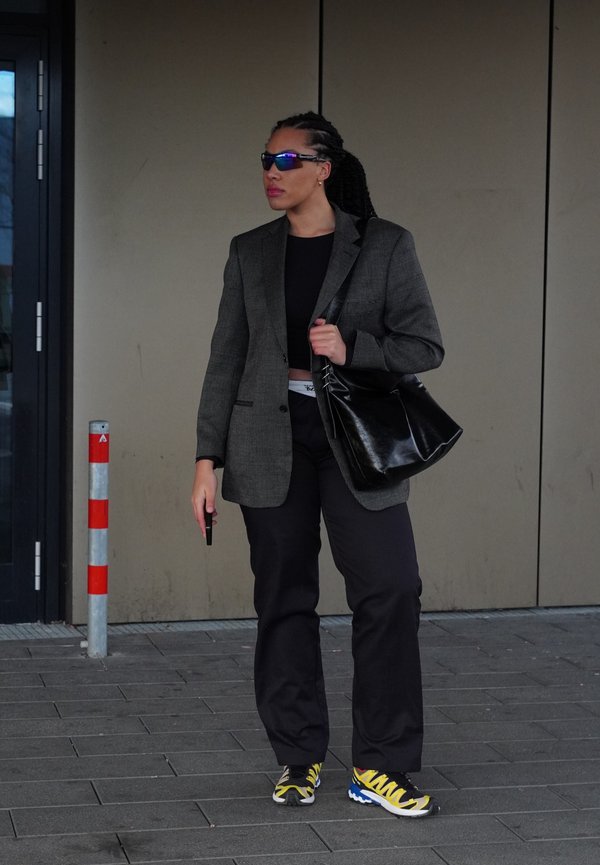 Žena nosi sivi sako, crni kratak top, crne hlače i tenisice s žutim akcentima, držeći crnu torbu i telefon.